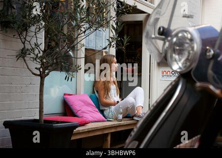Frau, die vor dem Amsterdamer Café sitzt, mit Vespa im Vordergrund Stockfoto