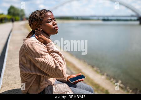 Die junge schöne schwarze Frau sitzt am Fluss in der Stadt. Sie hat Schmerzen im Nacken. Stockfoto