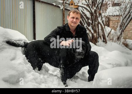 Ein Mann und ein schwarzer Labrador Retriever spielen im Neuschnee Stockfoto
