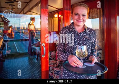 Kellnerin mit Glas Weißwein im Rovos Rail, Zug, von Pretoria nach Victoria Falls, Gauteng, Südafrika. Passagiere sitzen draußen im Freien Stockfoto