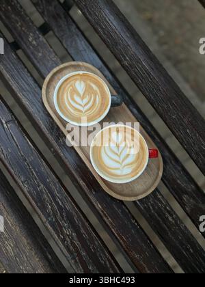 Zwei Cappuccinos mit Latte Art auf einem rustikalen Holztablett, serviert im Freien auf einer verwitterten Bank. Stockfoto
