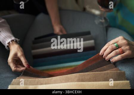 Zwei junge Erwachsene kaukasische Frauen wählen gemeinsam Stoffproben aus, Hände halten Farbmuster in verschiedenen Farben, eine Frau hält Weinglas und engagiert sich bei kreativen Entscheidungen Stockfoto