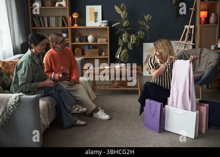 Drei kaukasische Frauen mittleren Alters sitzen zusammen im Wohnzimmer, lächeln und reden, während eine Frau neue Kleidung aus Einkaufstaschen zeigt, andere Weingläser in der Hand halten Stockfoto