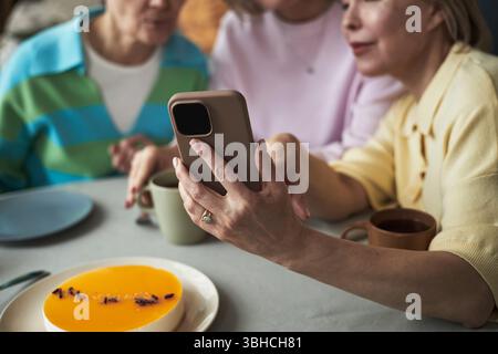 Drei kaukasische Frauen mittleren Alters sitzen zusammen am Tisch und machen Selfie mit dem Smartphone, lächeln und lehnen sich aus der Nähe, genießen Dessert und Getränke, Hände und Gesichter teilweise sichtbar Stockfoto