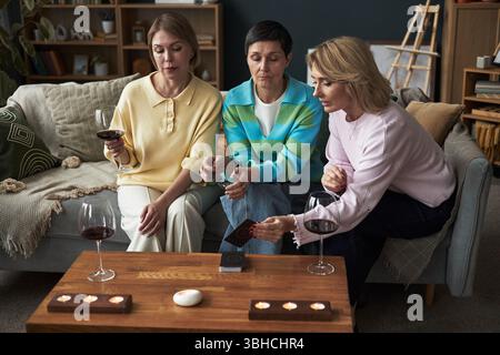 Drei weiße Frauen mittleren Alters sitzen auf dem Sofa und halten Gläser Rotwein, spielen gemeinsam Kartenspiel, unterhalten sich, Kerzen brennen auf Holztisch im Wohnzimmer Stockfoto