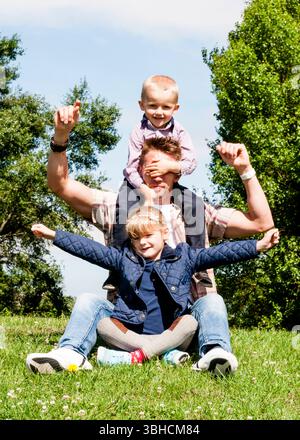 Ein Vater sitzt mit erhobenen Armen auf dem Gras, während seine Tochter im Kreuz auf seinem Schoß sitzt und sein Sohn auf seinen Schultern sitzt. Der Sohn hat einen Hektar Stockfoto