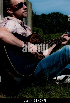 Ein Mann mit Sonnenbrille, kariertem Kurzarm-Hemd, blauer Jeans und weißen Sneakers sitzt vor einem Holzzaun und spielt eine schwarze Akustikgitarre. Er Stockfoto