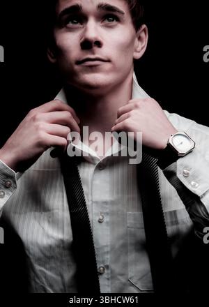 Ein junger Mann stellt seine schwarze Krawatte vor einen dunklen Hintergrund. Er trägt ein weißes Nadelstreifenhemd und eine Armbanduhr. Er schaut nach oben, Possi Stockfoto