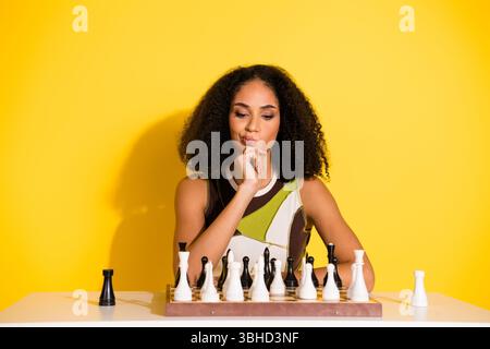Junge, stilvolle Frau, die sich auf die Entscheidung des Schachspiels konzentriert, vor gelbem Hintergrund, trendiges Outfit mit durchdachtem Ausdruck Stockfoto