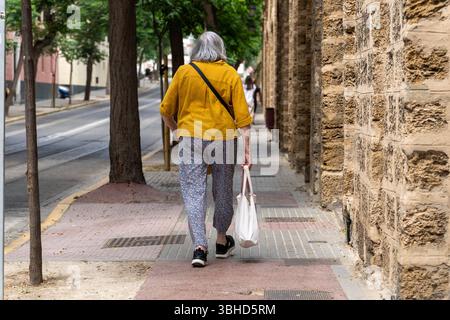 Cadiz, Andalusien, Spanien - 9. Juni 2025: Eine ältere Frau mit Einkaufstasche geht durch eine ruhige Straße in Cadiz. Symbolbild von Altersarmut, Inflation und Lebenswirklichkeit älterer Menschen *** eine ältere Frau geht mit Einkaufstasche durch eine ruhige Straße in Cadiz. Symbolbild Altersarmut, Inflation und die Lebensrealität älterer Menschen Stockfoto
