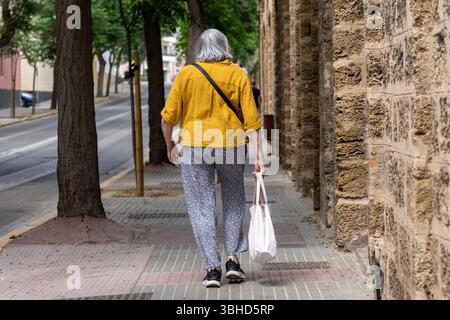 Cadiz, Andalusien, Spanien - 9. Juni 2025: Eine ältere Frau mit Einkaufstasche geht durch eine ruhige Straße in Cadiz. Symbolbild von Altersarmut, Inflation und Lebenswirklichkeit älterer Menschen *** eine ältere Frau geht mit Einkaufstasche durch eine ruhige Straße in Cadiz. Symbolbild Altersarmut, Inflation und die Lebensrealität älterer Menschen Stockfoto