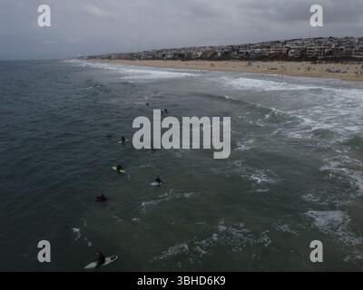 Aus der Vogelperspektive sehen Surfer, die in der Nähe des Manhattan Beach Pier unter stimmungsvollem Himmel Wellen fangen – Kaliforniens Küste in Bewegung von oben. Stockfoto