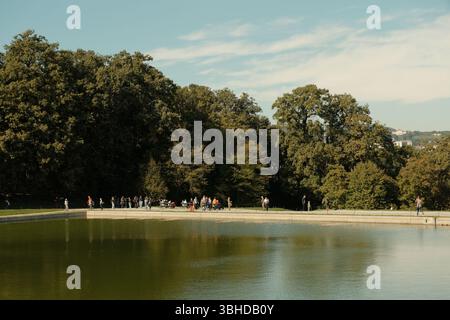 Eine ruhige Szene im Schönbrunner Garten, Wien, wo Besucher spazieren gehen und sich an einem reflektierenden Teich, der von üppigen Bäumen umgeben ist, versammeln. Stockfoto