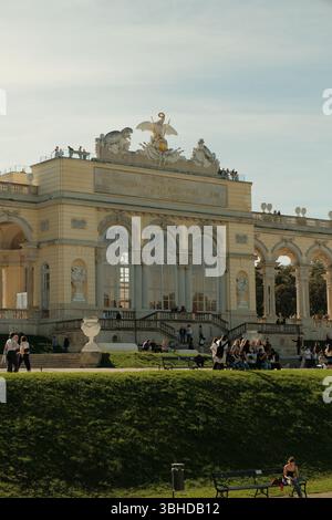 Die Gloriette im Schloss Schönbrunn in Wien ist von goldenem Sonnenlicht umgeben, umgeben von grünen Rasenflächen und Besuchergruppen, die den berühmten Barock genießen. Stockfoto