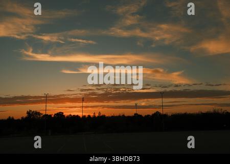 Dramatischer Sonnenuntergangshimmel mit orange, roten und goldenen Wolken über einem leeren Parkplatz. Hohe Lichtmasten bilden die Silhouette der Skyline. Stockfoto