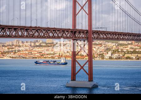 Farbenfrohe Landschaft mit blauem Containerschiff auf dem Tejo unter der berühmten Brücke 25 de Abril oder 25. April in Lissabon, Portugal. Berühmtes Eisenwerk der roten Hängebrücke, pulsierende Stadtlandschaft, städtische Architektur, Seeverkehr, sonniger Tag, bewölkter Himmel Stockfoto