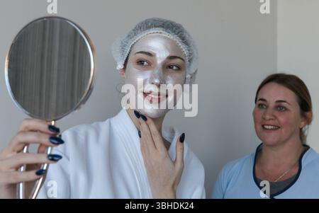 Junge Frau mit Gesichtsmaske lächelt, während sie im Spa-Salon auf einen Handspiegel blickt, Kosmetikerin steht hinter ihr und lächelt glücklich und zeigt eine Freude Stockfoto