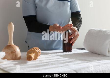 Weibliche Massagetherapeutin in professioneller Uniform Putting Lotion , und Organisieren von hölzernen Massagewerkzeugen, wie Rollen, auf einem Massagetisch in einer ruhigen B Stockfoto