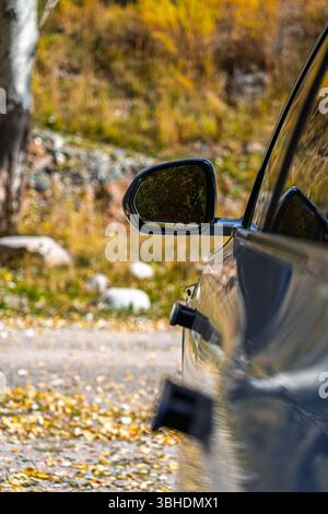 Bischkek, Kirgisistan - 12. Mai 2025: Nahaufnahme des Seitenspiegels eines dunklen Autos, der Bäume und Blätter im Herbst reflektiert, mit einem verschwommenen natürlichen Rücken Stockfoto