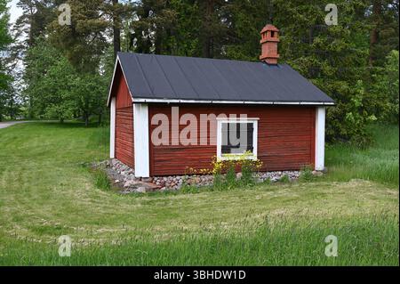 Ein charmantes traditionelles rotes Holzhaus in der finnischen Landschaft. Umgeben von üppigem grünem Gras und hohen Bäumen, hat die kleine Hütte einen Bla Stockfoto