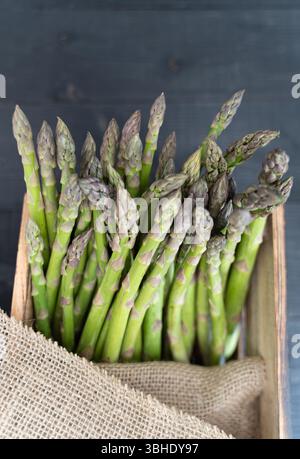 Top-down-Nahaufnahme von frischem grünem Spargel in einer Holzkiste, teilweise mit rustikalem Sacktuch umwickelt Stockfoto