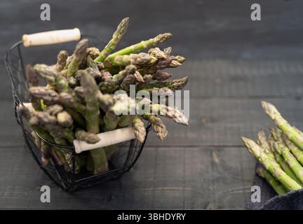 Top-down-Nahaufnahme von grünem Spargel im Metallkorb und rustikalem Stoffbündel auf dunklem Holztisch Stockfoto