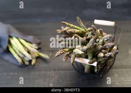Blick von oben auf frischen grünen Spargel in Drahtkorb mit einem in Küchentuch gewickelten Bündel auf schwarzer Holzoberfläche Stockfoto
