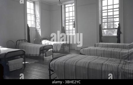 1950er Jahre, historisch, zahlreiche Einzelbetten in einem Schlafsaal in einem eleganten Zimmer mit hohen Schiebefenstern und Rollläden, Oxford, UK. Baumwollbettüberwürfe und individuelle Handtücher hängen am Ende der Metallbetten. Stockfoto