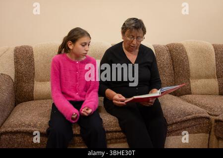 Eine Großmutter und Enkelin sitzen zusammen auf einer braun gestreiften Couch und verbringen viel Zeit damit, ein Buch zu lesen. Sie sind in legere Kleidung gekleidet Stockfoto