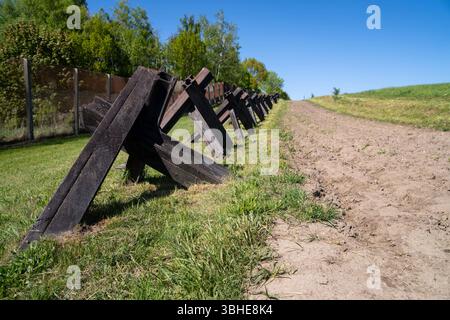 Hötensleben, Deutschland 27. April 2025: Im Bild liefert eine präzise Nahaufnahme der authentischen Grenzanlagen am Grenzdenkmal Hötensleben in Sachsen-Anhalt, einer der besterhaltenen Gedenkstätten der ehemaligen innerdeutschen Grenze. Im Vordergrund dominiert eine Reihe von Panzersperren, bekannt als Tschechenigel spanischer Reiter, die direkt neben dem Kolonnenweg positioniert sind. Der Kolonnenweg ist der unbefestigte Fahrweg, der von den Grenzsoldaten zur Patrouille und Grenzüberwachung genutzt wurde. Im Hintergrund ist ein Abschnitt eines inneren Grenzzauns oder einer Betonma Stockfoto