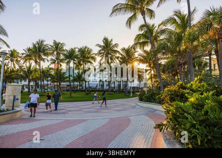 Abendlicher Blick auf die Fußgängerstraße in Miami Beach mit Touristen, die bei Sonnenuntergang unter Palmen in der Nähe von Art déco-Gebäuden spazieren. Miami Beach. USA. Stockfoto