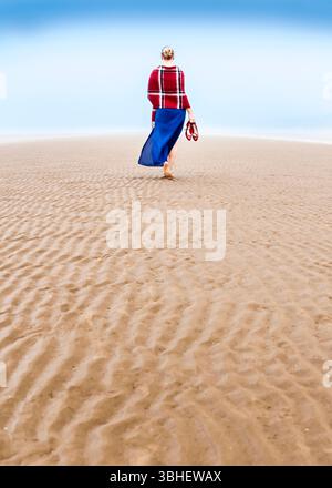 Eine Frau läuft barfuß über einen nassen Sandstrand und trägt ihre roten Schuhe in einer Hand. Sie trägt einen roten karierten Schal und einen langen blauen Rock. Der Strand Stockfoto