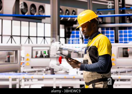 Der Mitarbeiter der automatisierten Fabrik verwaltet das industrielle Automatisierungssystem und gewährleistet Präzision in Förderband und Roboterarm. Der Techniker überprüft die Herstellung von Solarpaneelen mit Industrie 4,0-Software auf einem Tablet Stockfoto