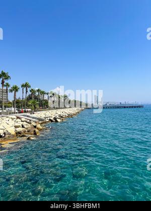 Städtische Küstenpromenade mit Palmen und kristallklarem türkisfarbenem Wasser an einem sonnigen Tag, mit modernen Wolkenkratzern und einem langen Pier im Hintergrund. Stockfoto