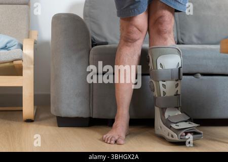 Nahaufnahme der Beine eines verletzten Mannes mit orthopädischem Laufstiefel, der in der Nähe des Sofas zu Hause steht, zeigt Erholung von Fuß- oder Knöchelverletzungen, Rehabilitationsprofi Stockfoto