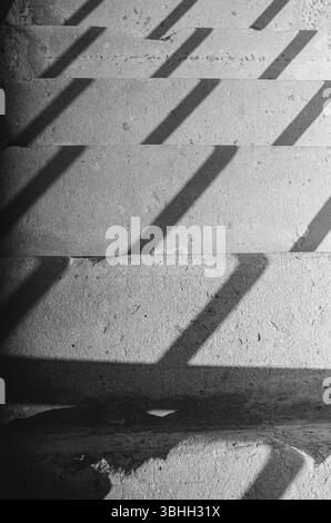 Schwarzweiß-Filmfoto mit diagonalen Schatten über abgenutzte Betontreppen, die Rhythmus, Textur und analoge Geometrie betonen. Stockfoto
