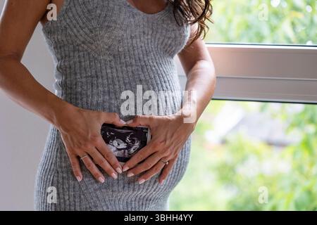 Großaufnahme der herzförmigen Hände der Frau, die Ultraschall ihres ersten Babys neben ihrem schwangeren Bauch hält Stockfoto