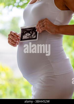 Nahaufnahme einer jungen Mutter, die einen Ultraschallscan ihres Babys mit beiden Händen vor dem schwangeren Magen hält. Sie ist bequem in ihren elastischen Klamotten Stockfoto