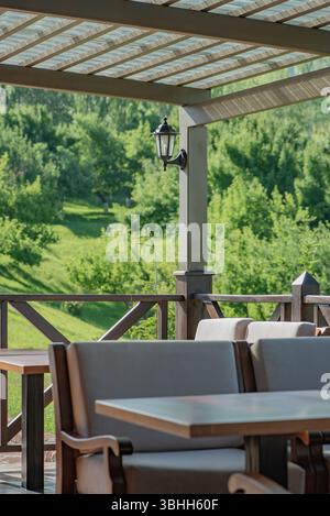 Eine gemütliche Terrasse mit weichen Stühlen und Holzmöbeln, einem Baldachin und einer Laterne vor einem grünen Hügel und dichtem Wald, der von hellem Sonnenlicht beleuchtet wird. Stockfoto