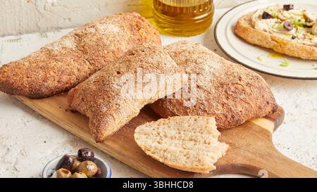 Dinkel-Ciabatta auf Holzoberfläche mit Oliven und Olivenöl im Hintergrund – ein rustikales Brot im mediterranen Stil, perfekt für Aperitif. Stockfoto