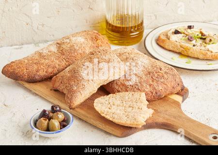 Dinkel-Ciabatta auf Holzoberfläche mit Oliven und Olivenöl im Hintergrund – ein rustikales Brot im mediterranen Stil, perfekt für Aperitif. Stockfoto
