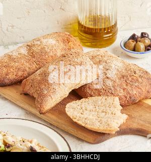 Dinkel-Ciabatta auf Holzoberfläche mit Oliven und Olivenöl im Hintergrund – ein rustikales Brot im mediterranen Stil, perfekt für Aperitif. Stockfoto