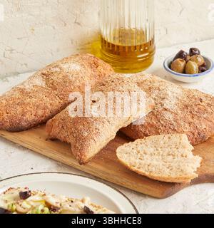 Dinkel-Ciabatta auf Holzoberfläche mit Oliven und Olivenöl im Hintergrund – ein rustikales Brot im mediterranen Stil, perfekt für Aperitif. Stockfoto