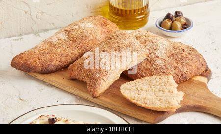 Dinkel-Ciabatta auf Holzoberfläche mit Oliven und Olivenöl im Hintergrund – ein rustikales Brot im mediterranen Stil, perfekt für Aperitif. Stockfoto
