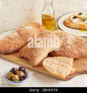 Dinkel-Ciabatta auf Holzoberfläche mit Oliven und Olivenöl im Hintergrund – ein rustikales Brot im mediterranen Stil, perfekt für Aperitif. Stockfoto