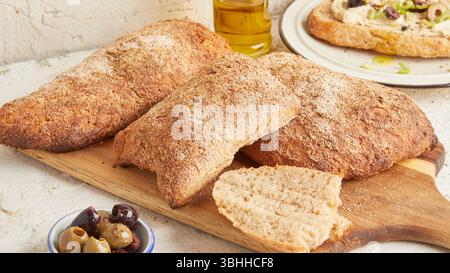 Dinkel-Ciabatta auf Holzoberfläche mit Oliven und Olivenöl im Hintergrund – ein rustikales Brot im mediterranen Stil, perfekt für Aperitif. Stockfoto