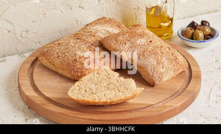 Dinkel-Ciabatta auf Holzoberfläche mit Oliven und Olivenöl im Hintergrund – ein rustikales Brot im mediterranen Stil, perfekt für Aperitif. Stockfoto