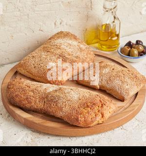 Dinkel-Ciabatta auf Holzoberfläche mit Oliven und Olivenöl im Hintergrund – ein rustikales Brot im mediterranen Stil, perfekt für Aperitif. Stockfoto