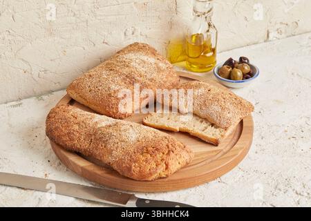 Dinkel-Ciabatta auf Holzoberfläche mit Oliven und Olivenöl im Hintergrund – ein rustikales Brot im mediterranen Stil, perfekt für Aperitif. Stockfoto