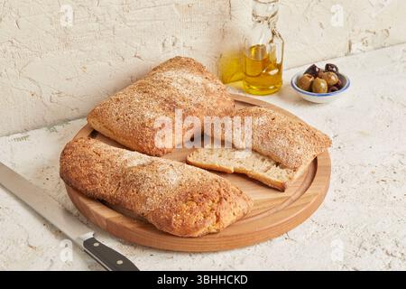 Dinkel-Ciabatta auf Holzoberfläche mit Oliven und Olivenöl im Hintergrund – ein rustikales Brot im mediterranen Stil, perfekt für Aperitif. Stockfoto